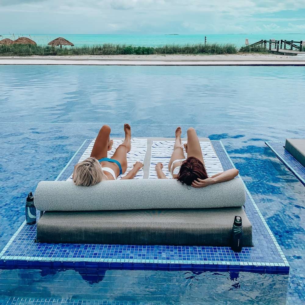 two women in bikinis lounge at oceanside pool with sandy beach in background