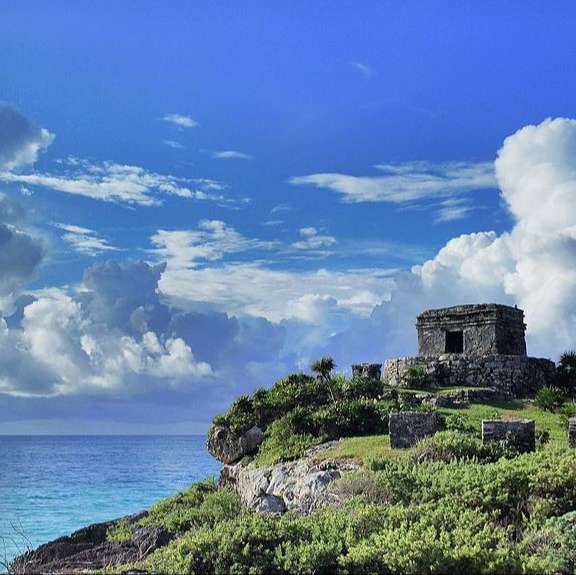 ancient stone temple on green cliff below large blue sky dotted with white clouds