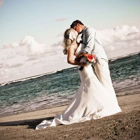 groom and bride both wear white and kiss oceanside